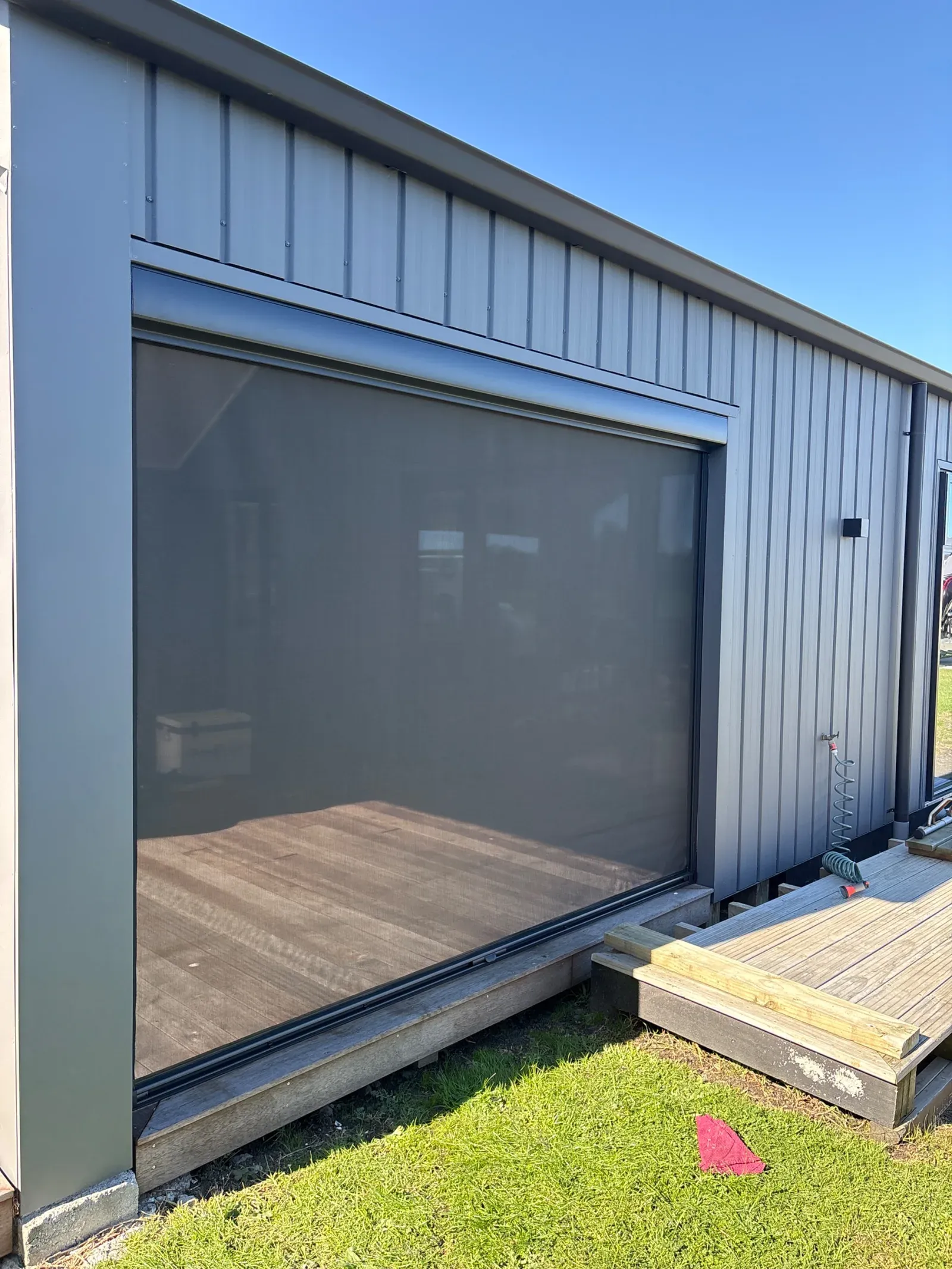 Charcoal Ziptrak outdoor blind lowered across a wide deck opening on a dark-clad house, seen from the lawn
