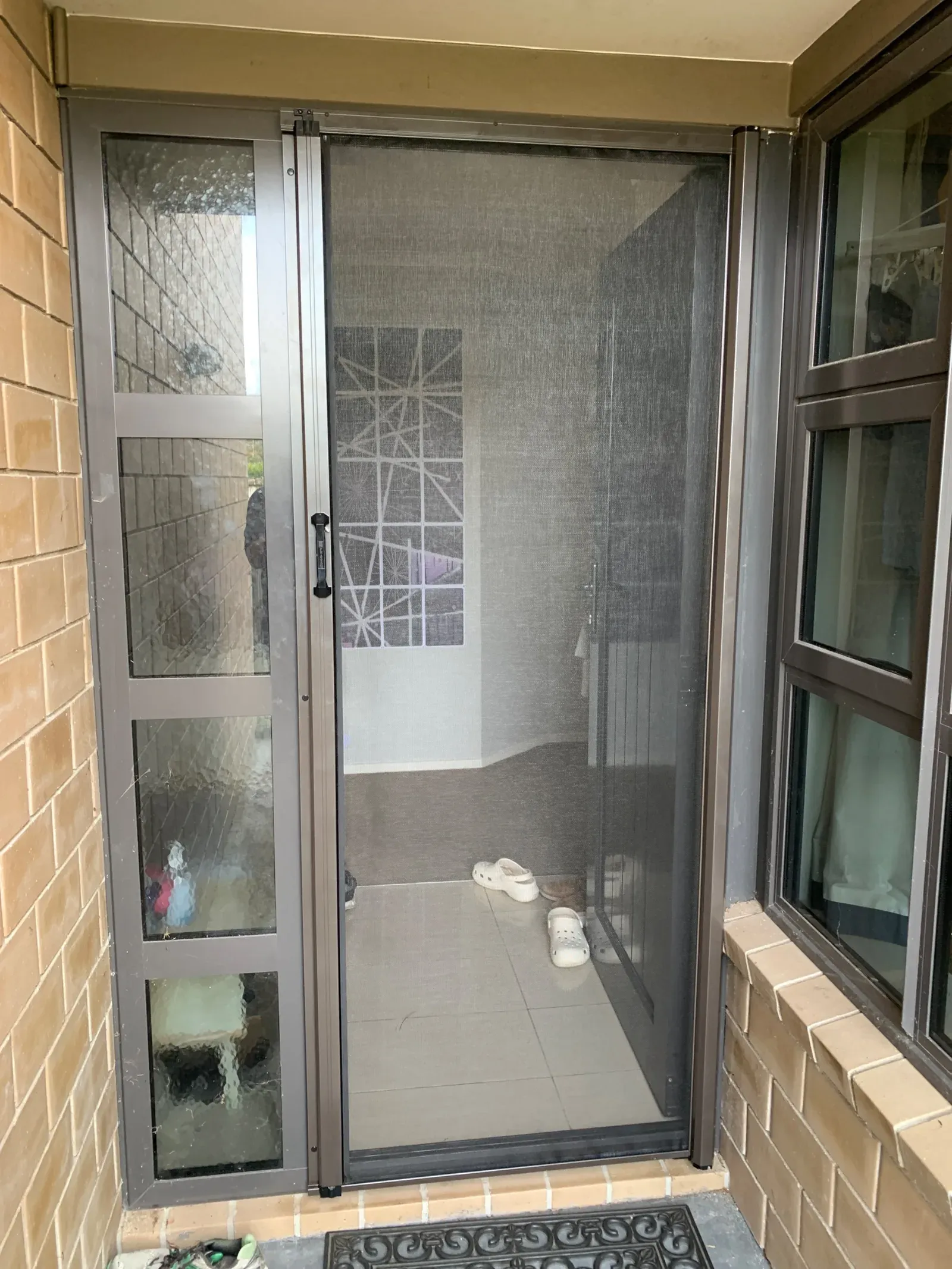 Bronze MeshLock retractable insect screen extended across a hinged entry door on a brick home, with a matching aluminium sidelight