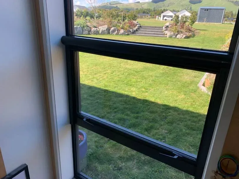 A black-framed awning window with a retractable insect screen, looking out over a rural lawn to green hills