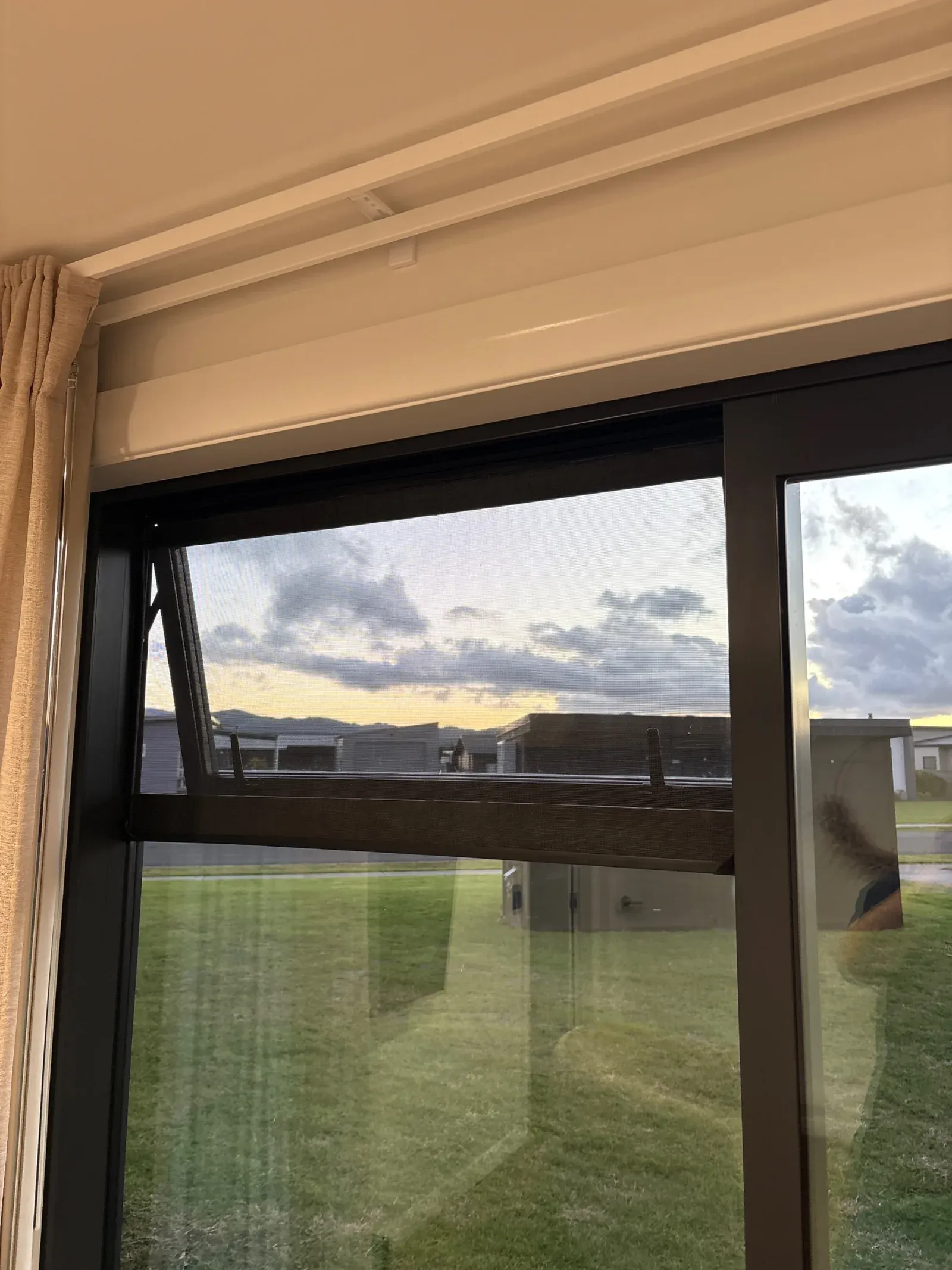 A black awning window fitted with a magnetic insect screen at dusk over a coastal lawn