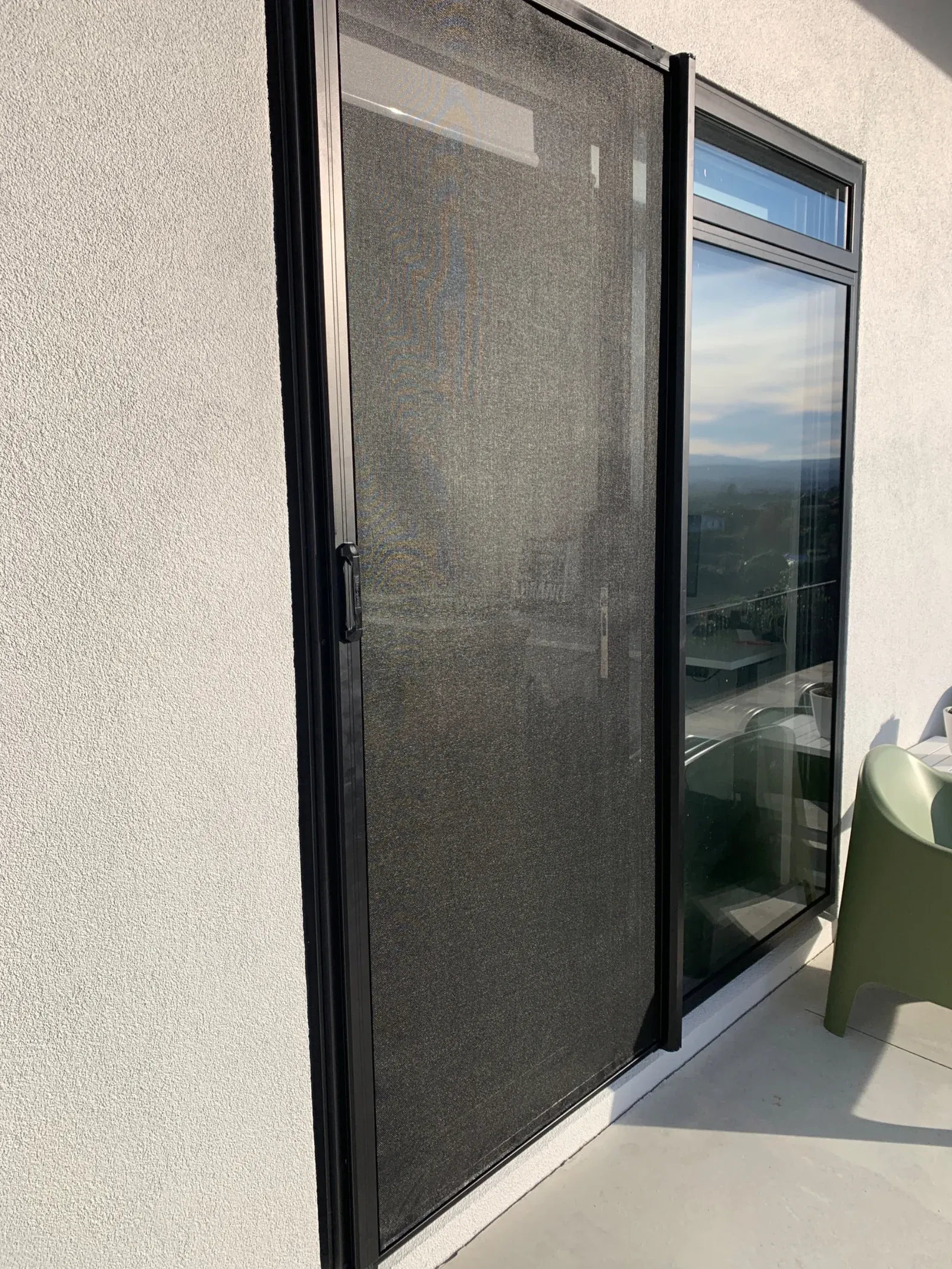 Black retractable insect screen door drawn across a sliding patio door on a sunny elevated deck, with a hillside view beyond