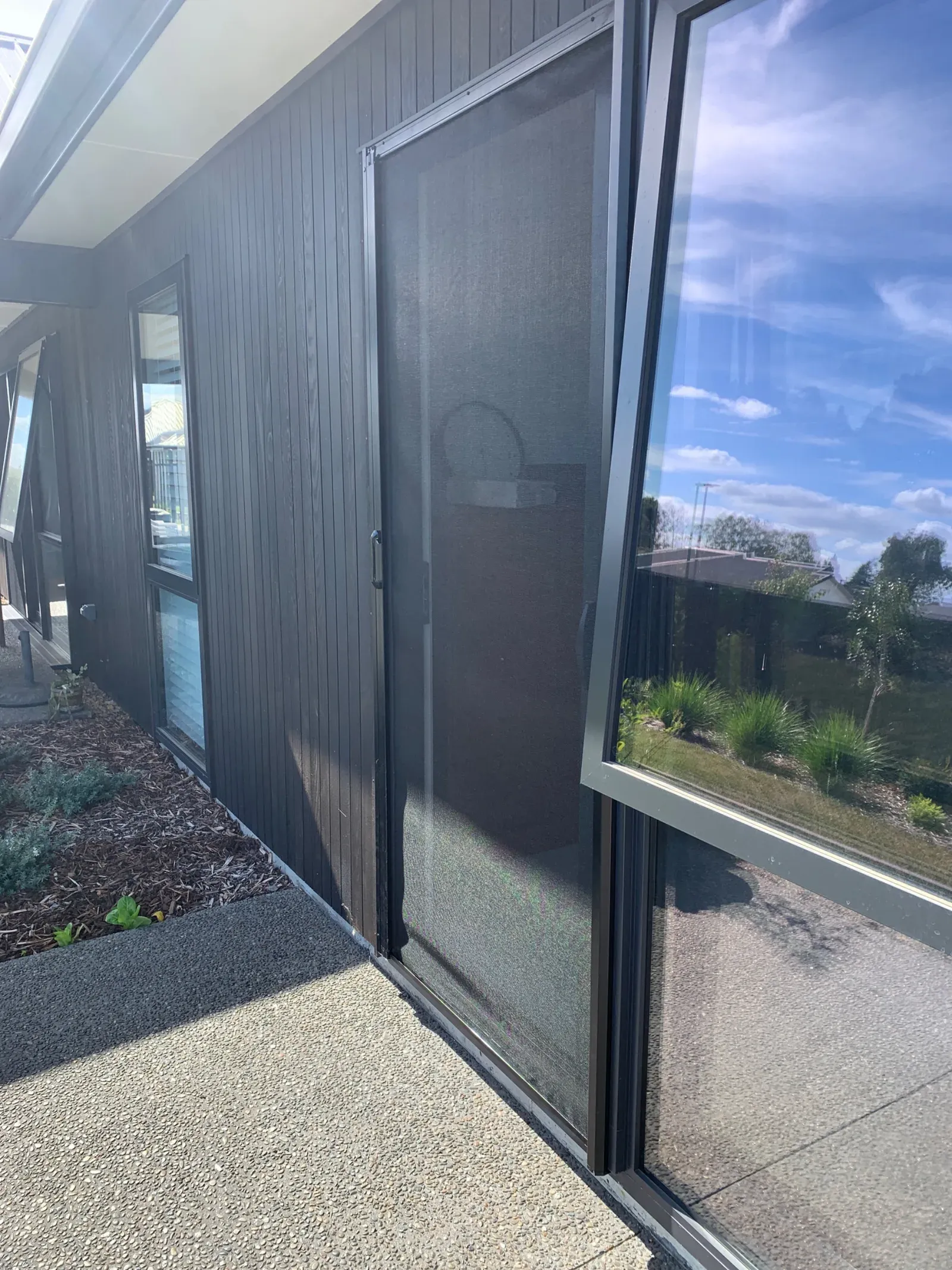 Charcoal retractable MeshLock screen drawn across a sliding door on a modern dark-weatherboard home under a blue sky