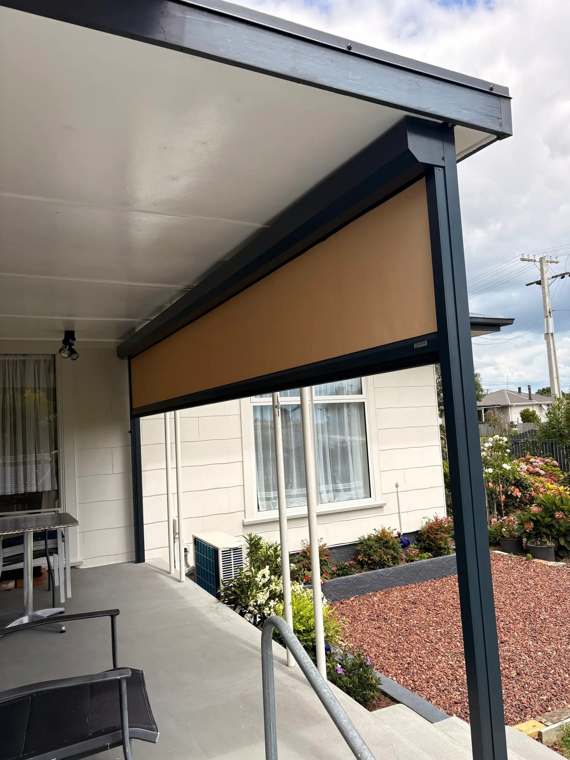 A tan outdoor blind part-lowered from its cassette under the verandah of a white weatherboard New Zealand home, with flowering garden beds alongside