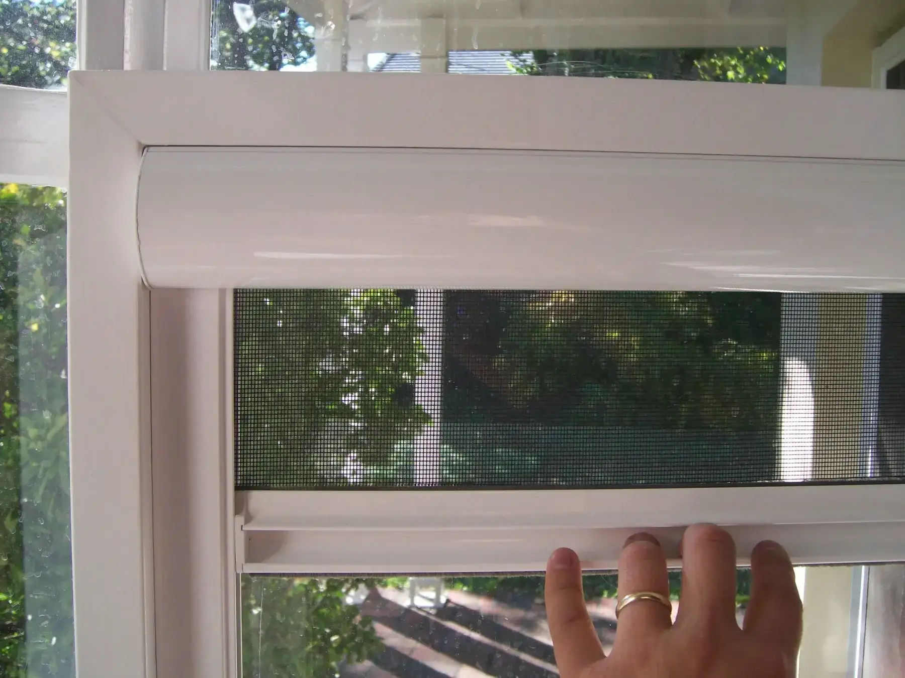 A hand guiding the mesh of a white retractable window insect screen down its side track