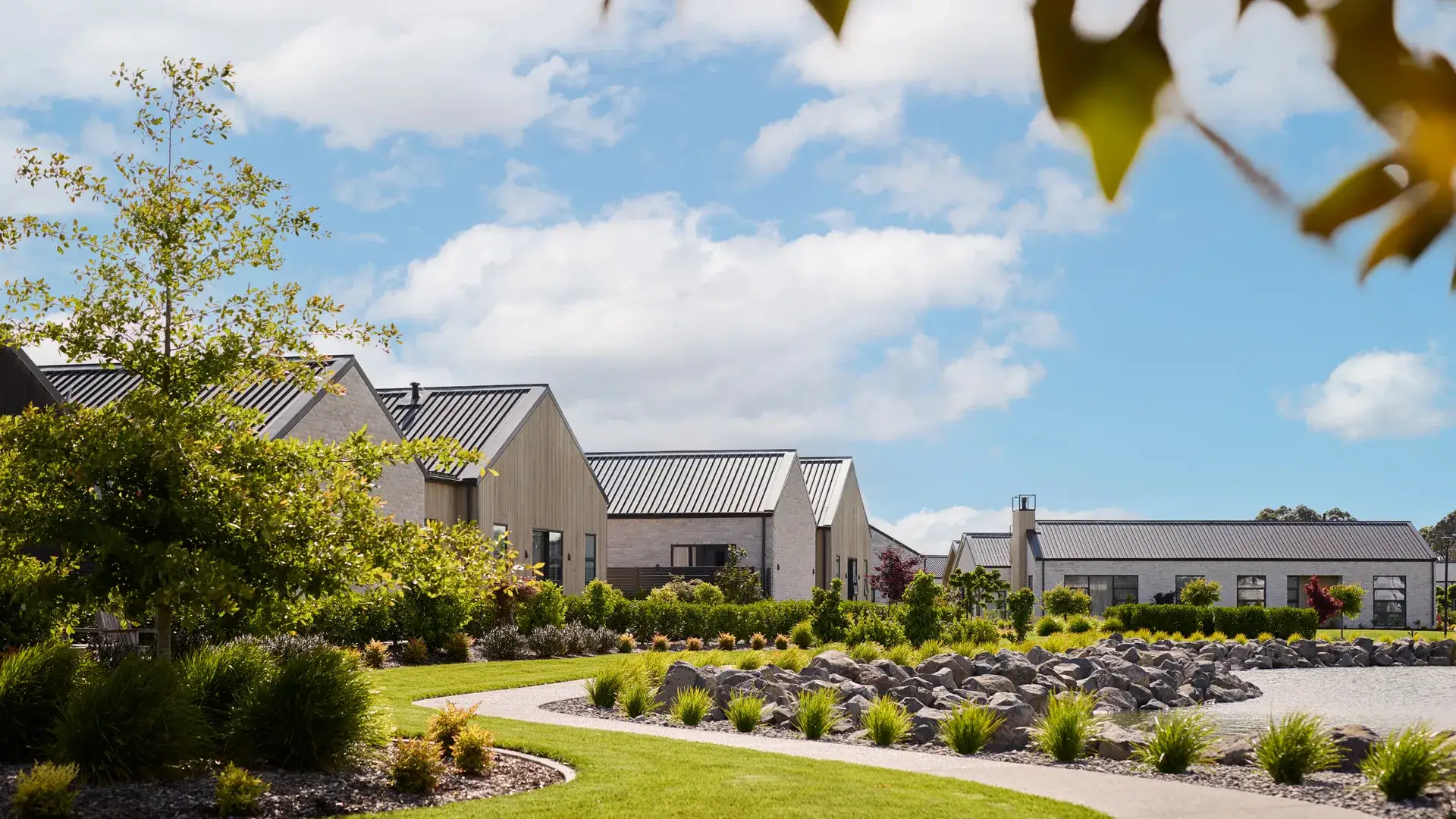 A landscaped retirement village of gable-roofed villas with a winding garden path, native plantings and a pond under a wide blue sky