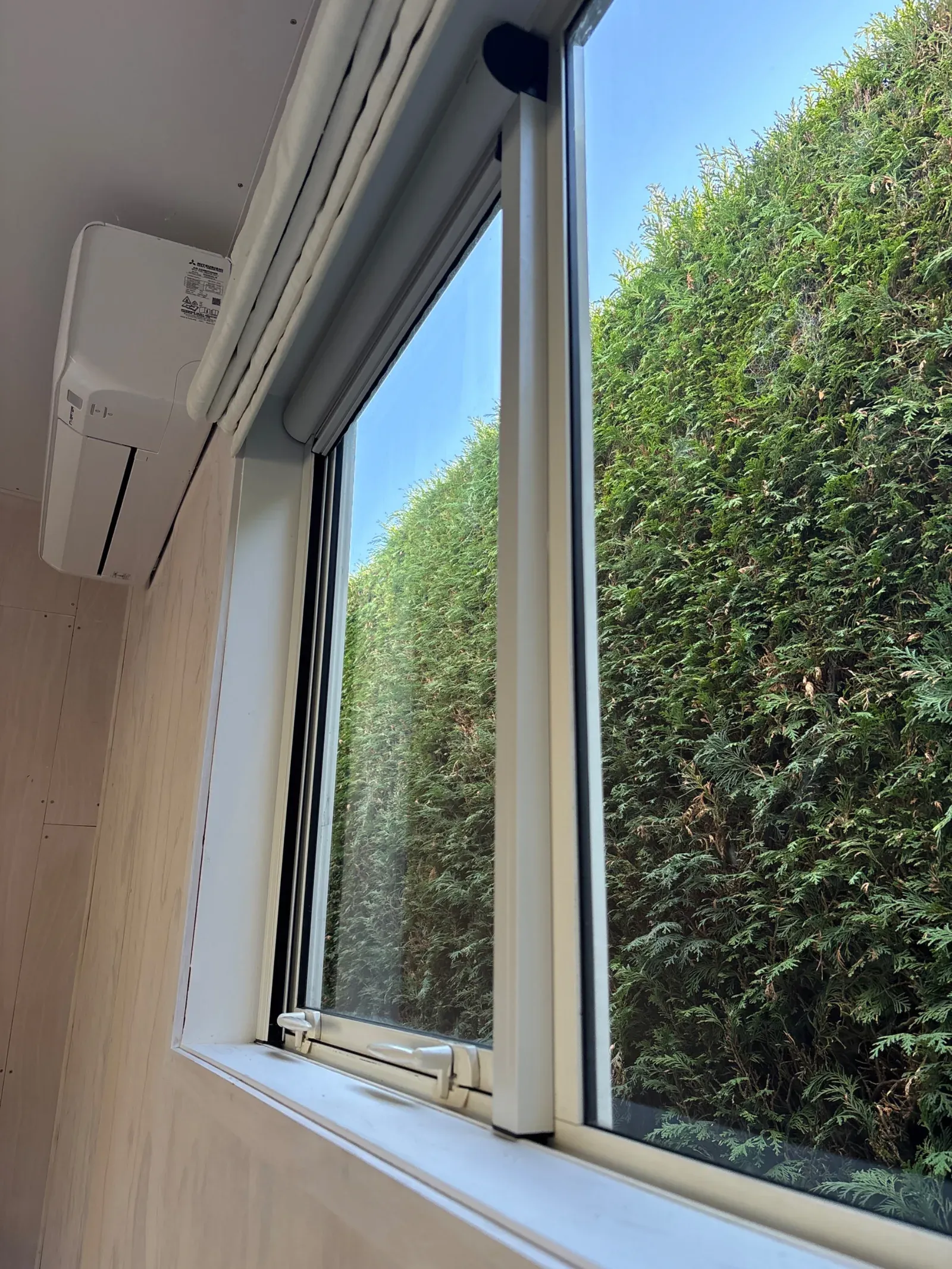 Retractable roller window screen fitted over an awning window, looking out to a tall green hedge, with a white cassette head-box above