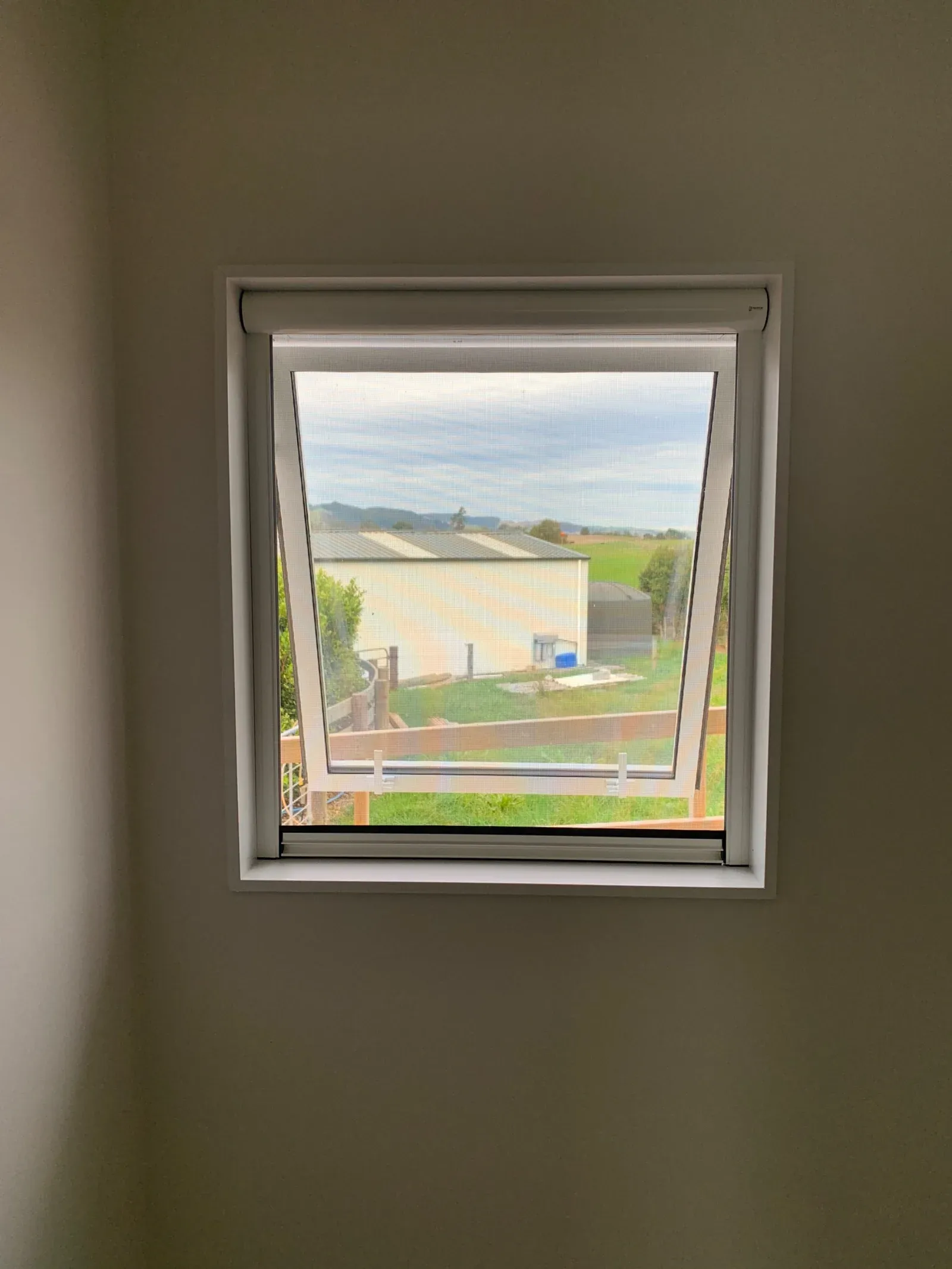 A retractable insect screen on a small awning window, the sash wound open over a farm shed and paddocks