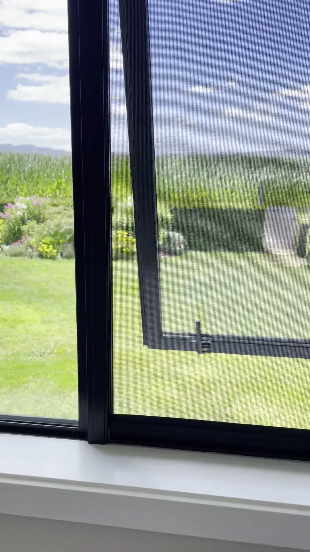 A black Serene insect screen drawn over an open awning window, garden and farmland beyond