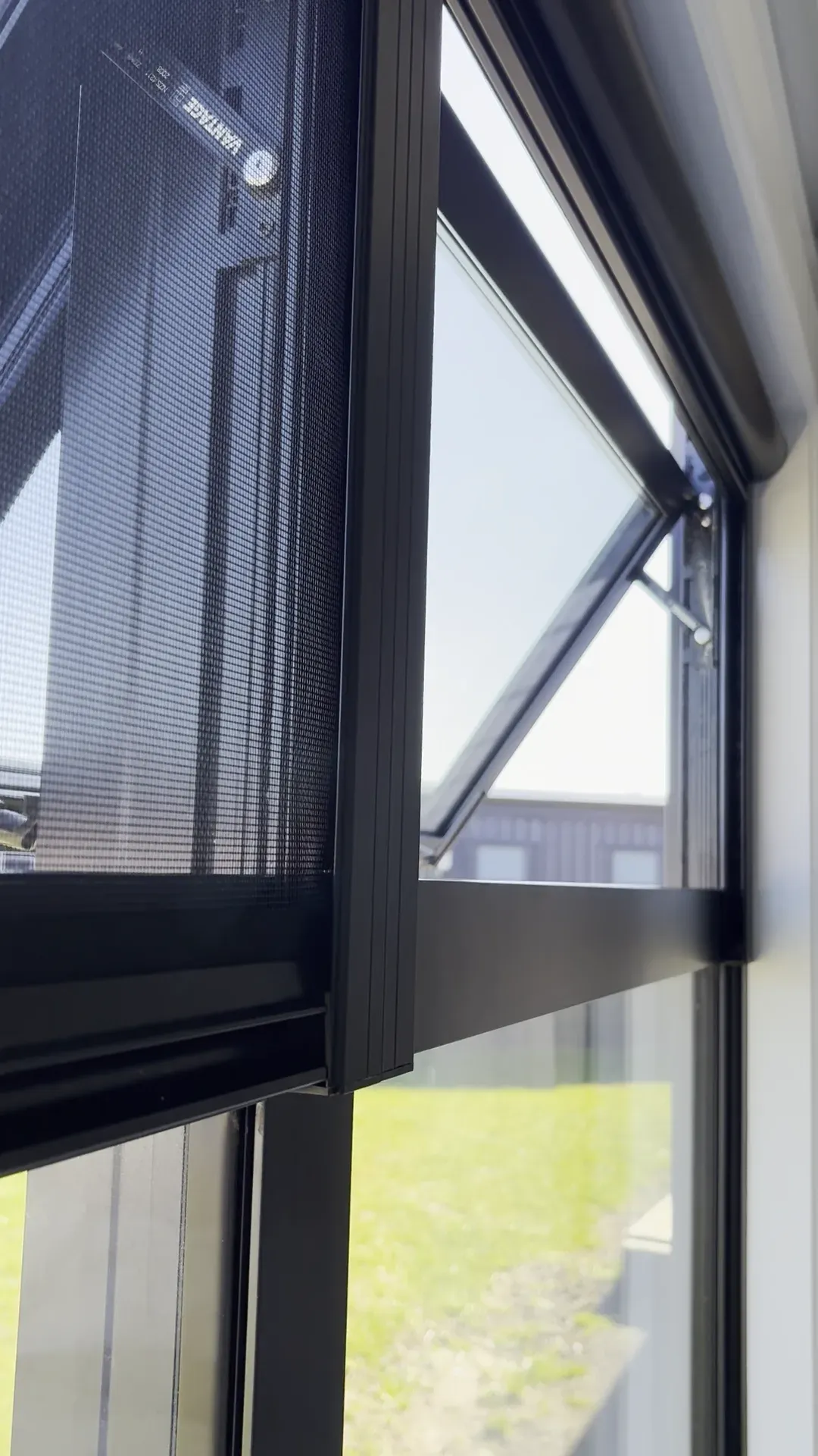 A black Serene insect screen drawn down over an open awning window, lawn and a coastal subdivision beyond